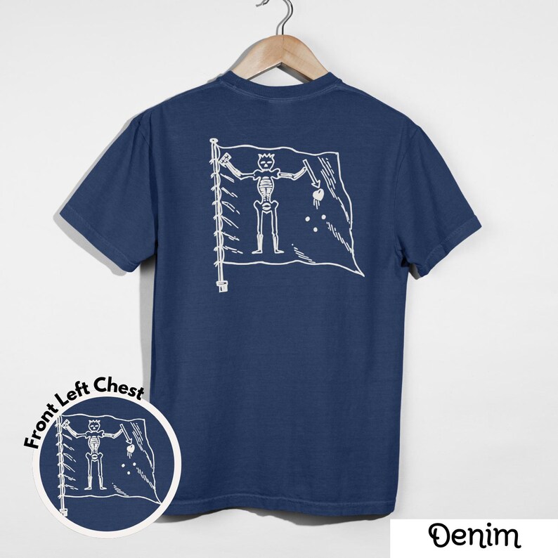 May include: Denim blue t-shirt with a white graphic of a skeleton on a flag. The flag design is on the back of the shirt. A smaller version of the graphic is on the front left chest. The word "Denim" is at the bottom right.