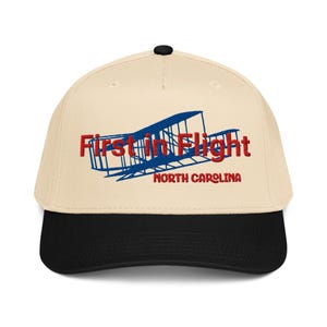 May include: A baseball cap with a cream-coloured crown and a black brim. The cap features the text "First in Flight" in red, with a blue airplane graphic, and "North Carolina" in red.