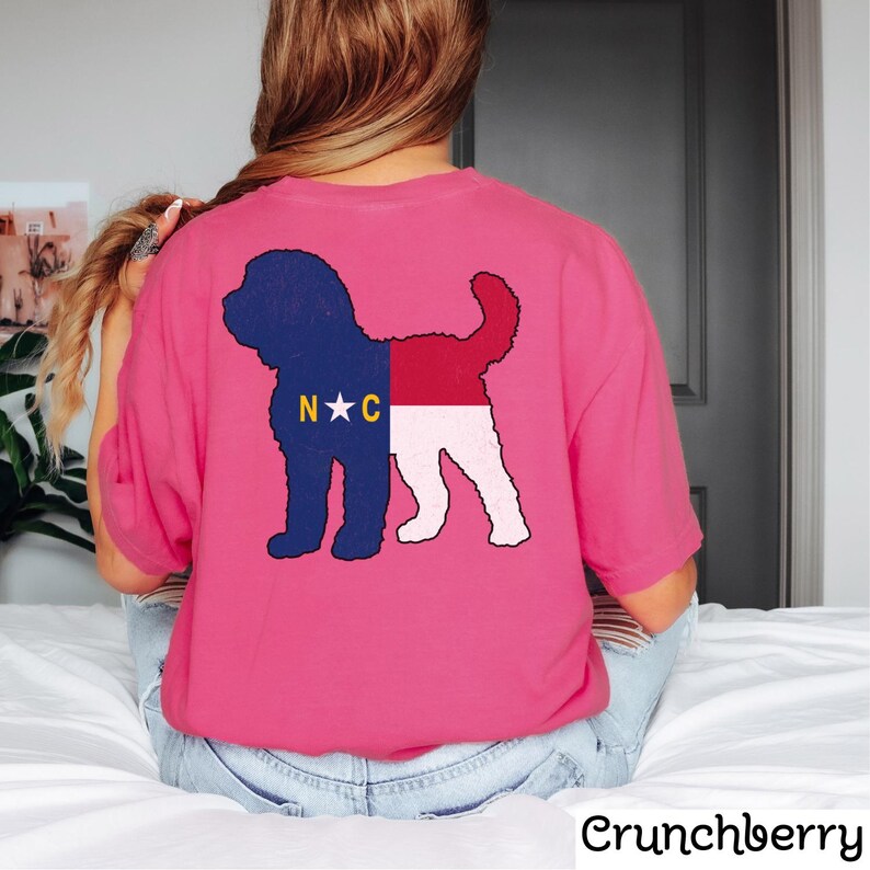 May include: Pink t-shirt featuring a silhouette of a dog with the North Carolina state flag design. The flag displays the letters "N.C." and a star in the centre.