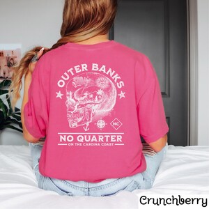 May include: Pink t-shirt with a white graphic of a skull with a compass and anchor. The text "Outer Banks No Quarter - On the Carolina Coast" is printed on the shirt.
