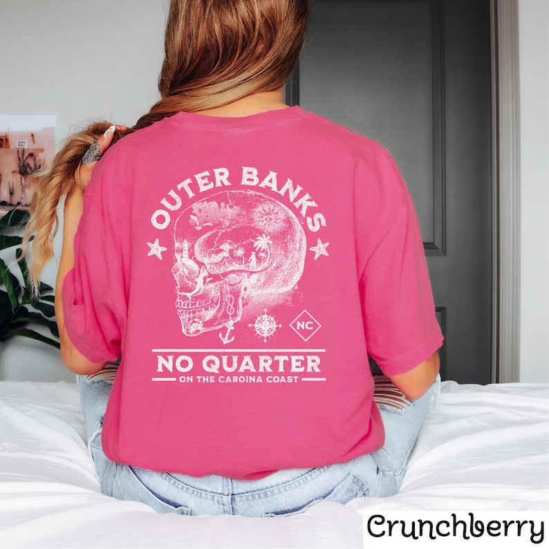 May include: Pink t-shirt with a white graphic of a skull with a compass and anchor. The text "Outer Banks No Quarter - On the Carolina Coast" is printed on the shirt.