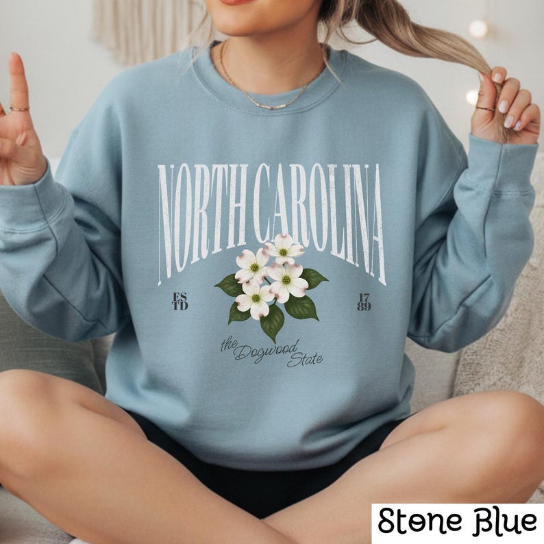May include: Stone blue sweatshirt with the text "NORTH CAROLINA" in white arched lettering. Below the text is a floral design with white flowers and green leaves. The text "the Dogwood State" is below the floral design. The sweatshirt has long sleeves.