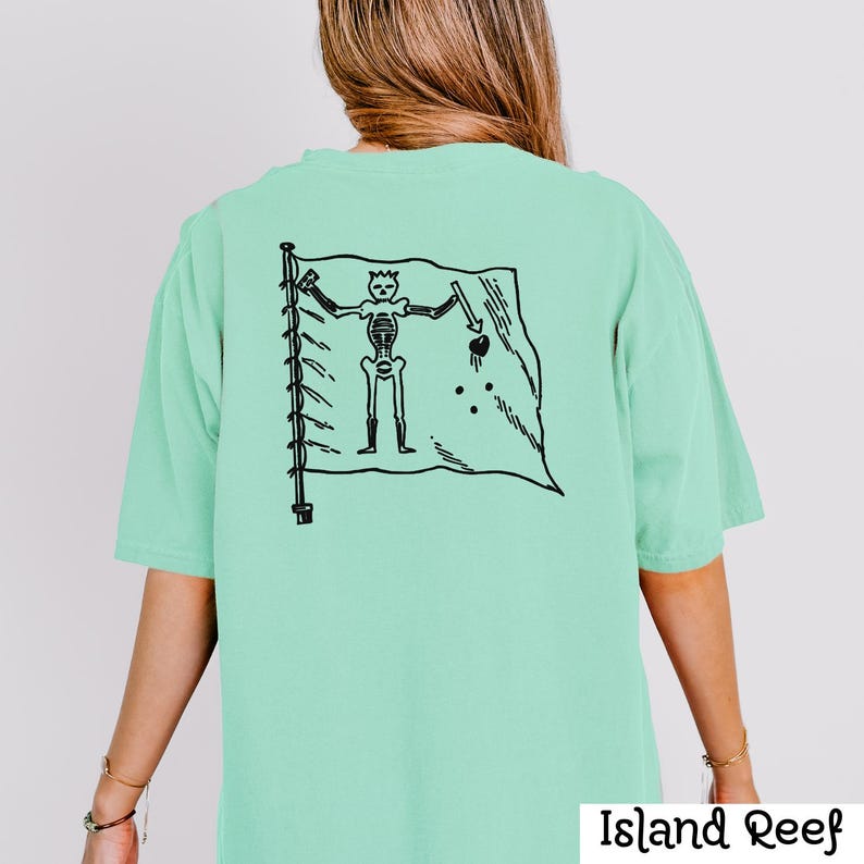 May include: A light green t-shirt with a black graphic of a skeleton pirate flag on the back. The flag features a skeleton holding a sword and a heart. The text "Island Reef" is at the bottom right.