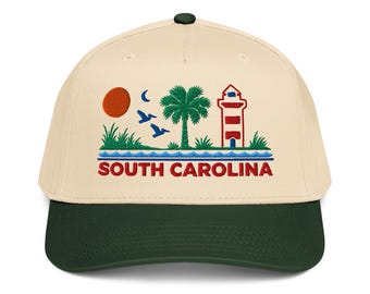 May include: A cream-colored baseball cap with a dark green brim and a top button. The front features an embroidered design of a red sun, a blue crescent moon, two flying pelicans, a green palm tree, a red and white lighthouse, and the text SOUTH CAROLINA.