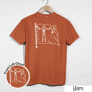 May include: A burnt orange t-shirt with a white graphic of a skeleton holding a sword and a heart on a flag. The shirt is hanging on a wooden hanger against a white background. The text "Yam" is at the bottom.