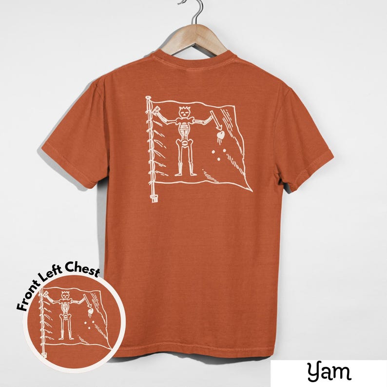 May include: A burnt orange t-shirt with a white graphic of a skeleton holding a sword and a heart on a flag. The shirt is hanging on a wooden hanger against a white background. The text "Yam" is at the bottom.