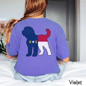 May include: A purple t-shirt featuring a silhouette of a dog outlined in the North Carolina state flag. The dog is facing right and the flag is red, white and blue with the letters "N.C." and a white star in the centre.