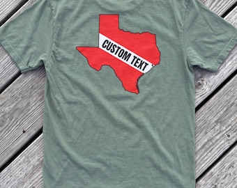 May include: A sage green short-sleeved t-shirt featuring a red graphic of the state of Texas on the back. A white diagonal stripe runs across the center of the state shape, containing the black text CUSTOM TEXT in a bold, sans-serif font.
