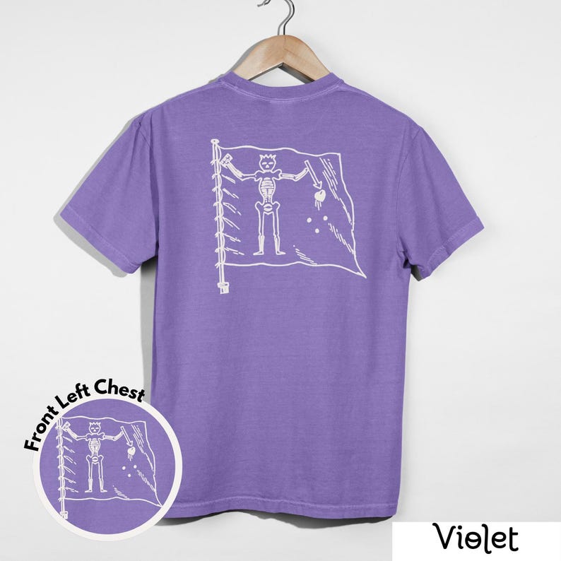 May include: Violet-colored t-shirt with a white graphic of a skeleton on a flag. The shirt is hanging on a wooden hanger. A circular graphic on the front left chest mirrors the design on the back. The word "Violet" is at the bottom right.
