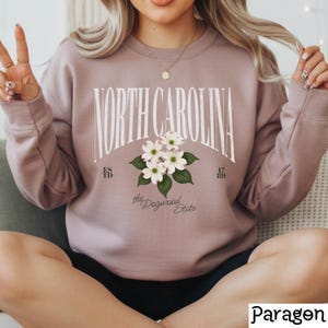 May include: A mauve sweatshirt with the text "NORTH CAROLINA" in a white, arched font. Below the text is a floral design with white flowers and green leaves. The words "the Dogwood State" are below the floral design.