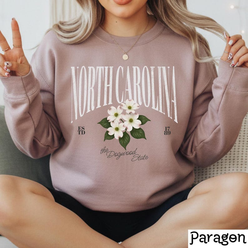 May include: A mauve sweatshirt with the text "NORTH CAROLINA" in a white, arched font. Below the text is a floral design with white flowers and green leaves. The words "the Dogwood State" are below the floral design.