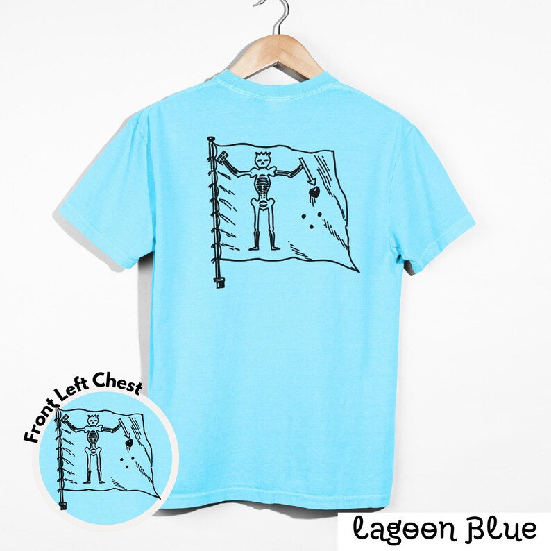 May include: A light blue t-shirt with a black graphic of a skeleton pirate flag on the back. The flag features a skeleton holding a sword and a heart. The words "Lagoon Blue" are printed at the bottom right.