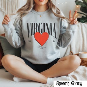 May include: Sport grey sweatshirt featuring the word "VIRGINIA" in black, a red heart, and the text "For Lovers." The sweatshirt also has the numbers "17" and "88" on either side of the heart. The sweatshirt is a crew neck.