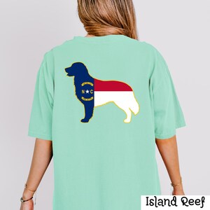 Puede incluir: Una camiseta verde claro con la silueta de un perro rellena con el dise&ntilde;o de la bandera del estado de Carolina del Norte. El contorno del perro es azul marino, con los colores de la bandera rojo, blanco y azul. El texto "Island Reef" est&aacute; en la parte inferior.