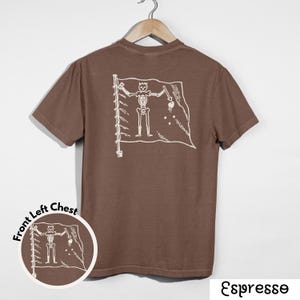 May include: Espresso-colored t-shirt with a white graphic of a skeleton holding a sword and a heart on a flag. The shirt is hanging on a wooden hanger. A smaller version of the graphic is on the front left chest.