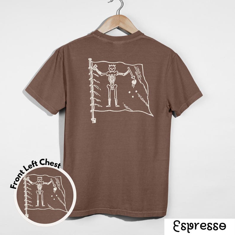 May include: Espresso-colored t-shirt with a white graphic of a skeleton holding a sword and a heart on a flag. The shirt is hanging on a wooden hanger. A smaller version of the graphic is on the front left chest.