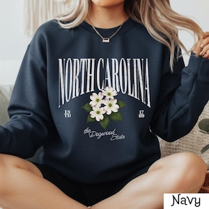 May include: Navy blue sweatshirt with the text "NORTH CAROLINA" in a white arched font, with a floral design of white dogwood blossoms. The text "ESTD 1789" and "the Dogwood State" are also present.