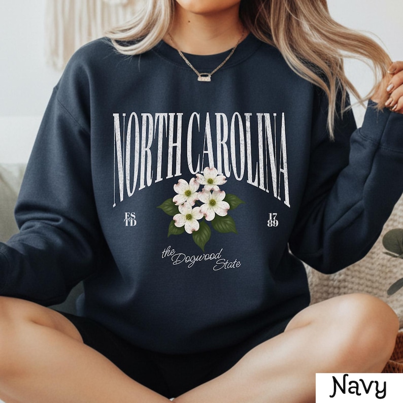 May include: Navy blue sweatshirt with the text "NORTH CAROLINA" in a white arched font, with a floral design of white dogwood blossoms. The text "ESTD 1789" and "the Dogwood State" are also present.