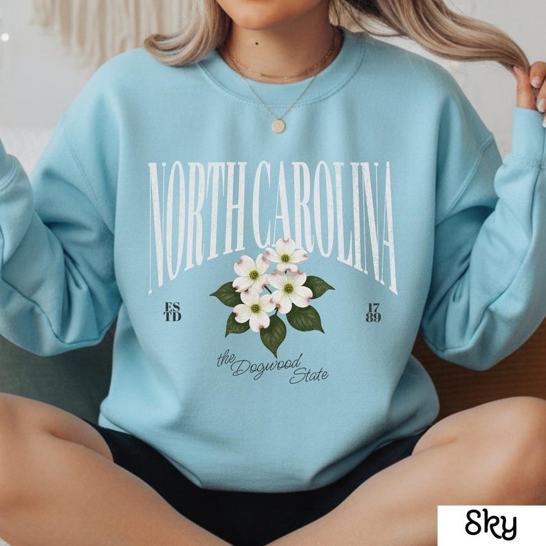 May include: Sky blue sweatshirt with the words "NORTH CAROLINA" in white block letters. Below the text is a floral design of white dogwood blossoms and green leaves. The sweatshirt also has the text "the Dogwood State", "ESTD" and "17 89".