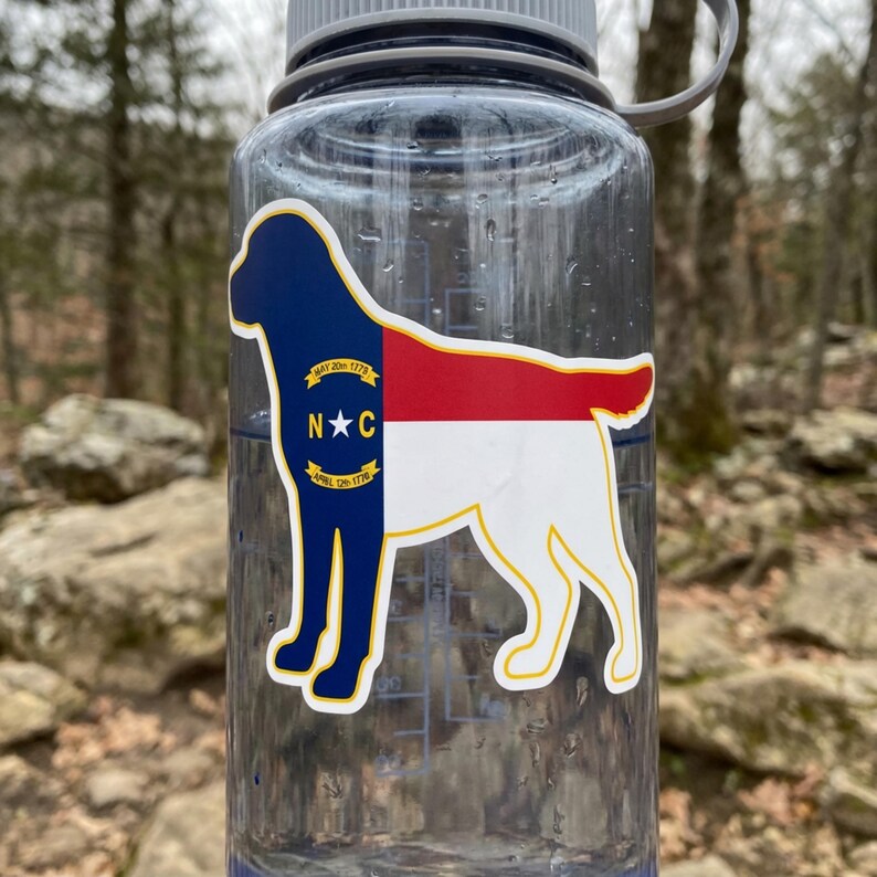 May include: A clear water bottle with a sticker featuring the North Carolina state flag in the shape of a dog. The sticker has a blue section with text, a red section, and a white section. The bottle is outdoors.