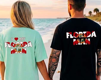 May include: A light mint green t-shirt with the text I heart MY FLORIDA MAN and a black t-shirt with the text FLORIDA MAN, both featuring the Florida state flag design within the letters, displayed on the back of two shirts.