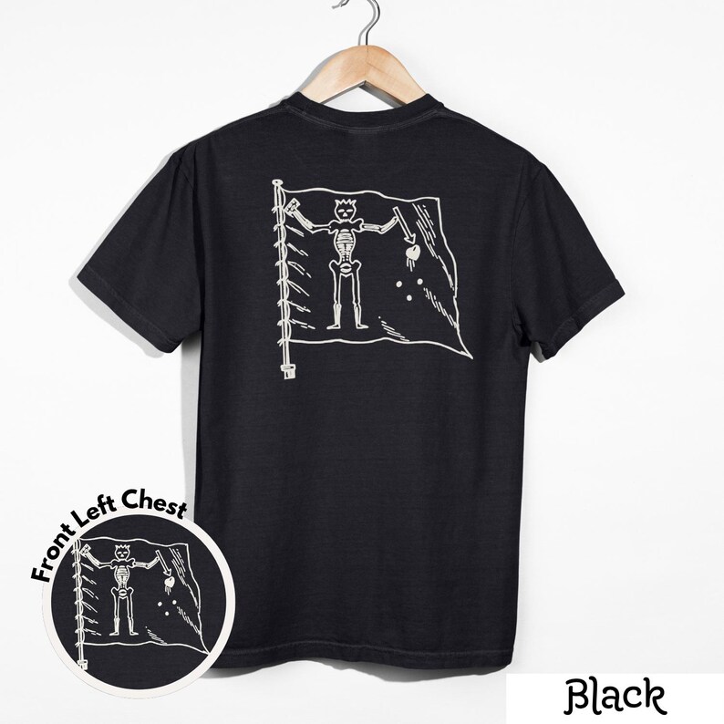 May include: Black t-shirt featuring a white graphic of a pirate flag with a skeleton figure. The flag design includes a skull, crossbones, and other symbols. The shirt is displayed on a wooden hanger.