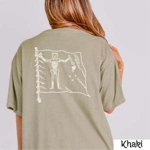 May include: Khaki-colored t-shirt featuring a white graphic of a pirate flag on the back. The flag design includes a skeleton figure and other pirate-themed elements. The shirt is a casual, oversized style.