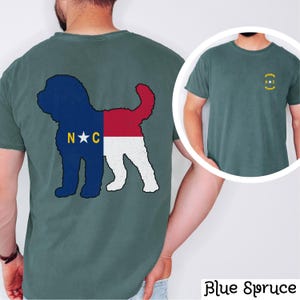 May include: A teal blue t-shirt featuring a silhouette of a dog with the North Carolina state flag design. The back of the shirt displays the silhouette of the dog with the flag design. The front of the shirt has a small circle containing the letters "N.C."