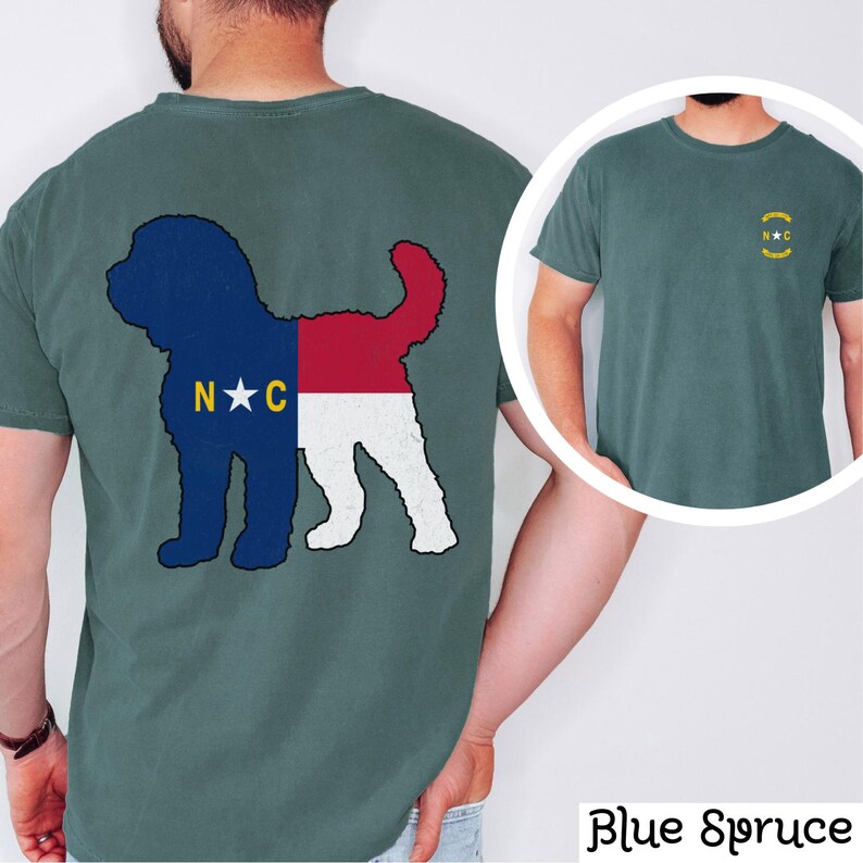 May include: A teal blue t-shirt featuring a silhouette of a dog with the North Carolina state flag design. The back of the shirt displays the silhouette of the dog with the flag design. The front of the shirt has a small circle containing the letters "N.C."