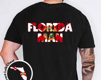 May include: A black short-sleeved t-shirt featuring the text FLORIDA MAN printed in white with a red cross and the Florida state seal pattern inside the letters. A circular inset shows a white outline of Florida with the state seal on the front left chest.