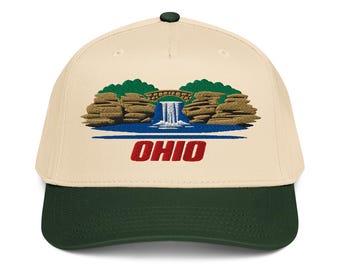 May include: A cream-colored baseball cap with a dark green brim and a top button. The front features an embroidered graphic of a waterfall flowing under a stone bridge between rocky cliffs, with the word OHIO in red letters underneath the water.