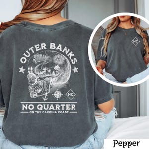 May include: A dark gray t-shirt with a white graphic on the back. The graphic features a skull with a compass and the text "Outer Banks No Quarter - On the Carolina Coast". The front of the shirt has a white "NC" in a square on the left chest.