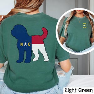May include: A light green t-shirt featuring a silhouette of a dog with the North Carolina flag design on the back. The front of the shirt has a small North Carolina flag design with the letters "N.C." in a circle.