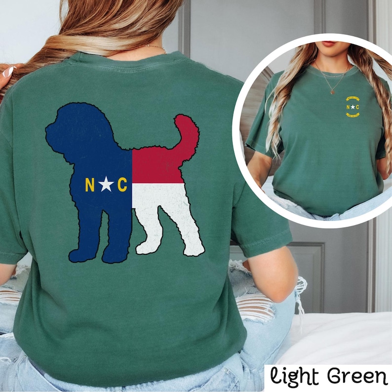 May include: A light green t-shirt featuring a silhouette of a dog with the North Carolina flag design on the back. The front of the shirt has a small North Carolina flag design with the letters "N.C." in a circle.