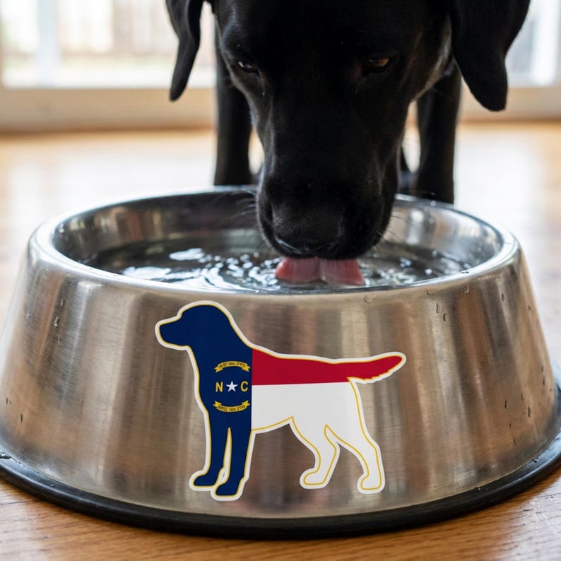May include: A stainless steel dog bowl with a sticker of a dog in the North Carolina state flag design. A black dog is drinking water from the bowl. The sticker features the state's colors: blue, red, and white.
