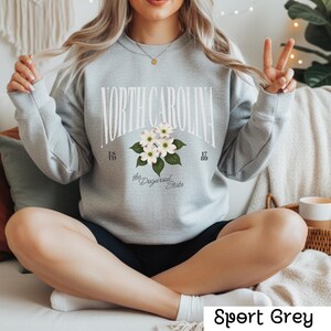 May include: Sport Grey sweatshirt featuring the text "NORTH CAROLINA" in a curved arch, with a floral design of white dogwood blossoms. The sweatshirt also includes the text "the Dogwood State" and the years "1789" and "1815".