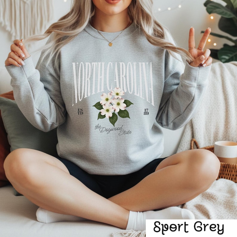May include: Sport Grey sweatshirt featuring the text "NORTH CAROLINA" in a curved arch, with a floral design of white dogwood blossoms. The sweatshirt also includes the text "the Dogwood State" and the years "1789" and "1815".