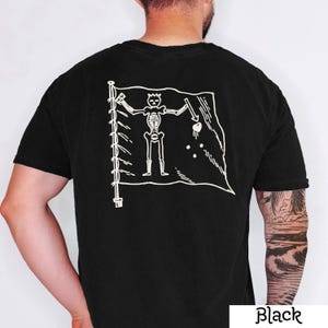 May include: Black t-shirt featuring a white graphic of a pirate flag with a skeleton figure holding a spear and a heart. The flag is on a pole with a series of white lines. The word "Black" is printed in white at the bottom right.