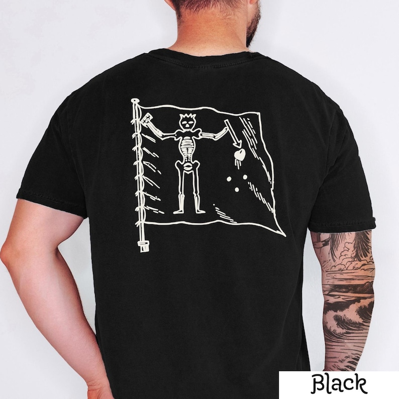 May include: Black t-shirt featuring a white graphic of a pirate flag with a skeleton figure holding a spear and a heart. The flag is on a pole with a series of white lines. The word "Black" is printed in white at the bottom right.