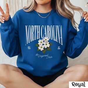 May include: Royal blue sweatshirt with the text "NORTH CAROLINA" in a white arched font. Below the text is a floral design of white dogwood blossoms and green leaves. The sweatshirt also has the text "the Dogwood State".