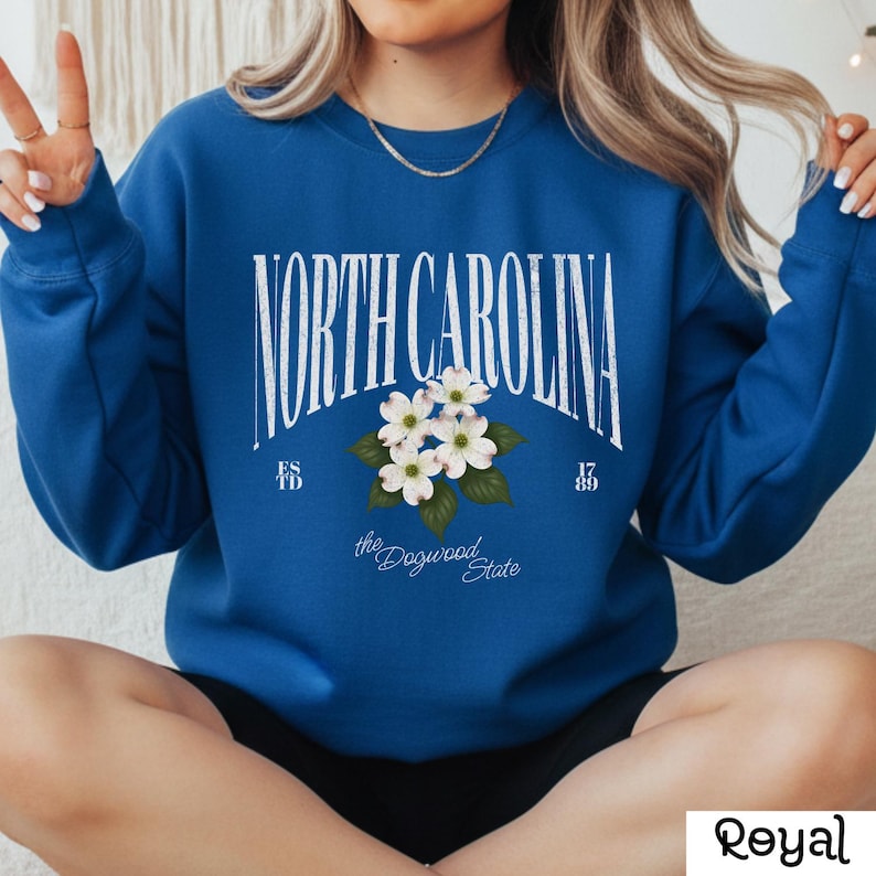 May include: Royal blue sweatshirt with the text "NORTH CAROLINA" in a white arched font. Below the text is a floral design of white dogwood blossoms and green leaves. The sweatshirt also has the text "the Dogwood State".