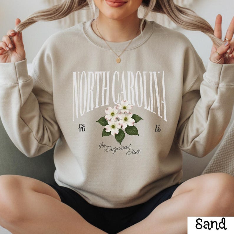 May include: Sand-colored sweatshirt with the text "NORTH CAROLINA" in white arched lettering. Below the text is a floral design with white flowers and green leaves. The words "the Dogwood State" are below the floral design.