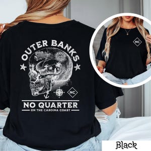 May include: Black t-shirt with a white graphic of a skull with a compass and anchor. The text "Outer Banks No Quarter - On the Carolina Coast" is printed on the shirt. The shirt also has a small white square with the letters "NC" on the back.