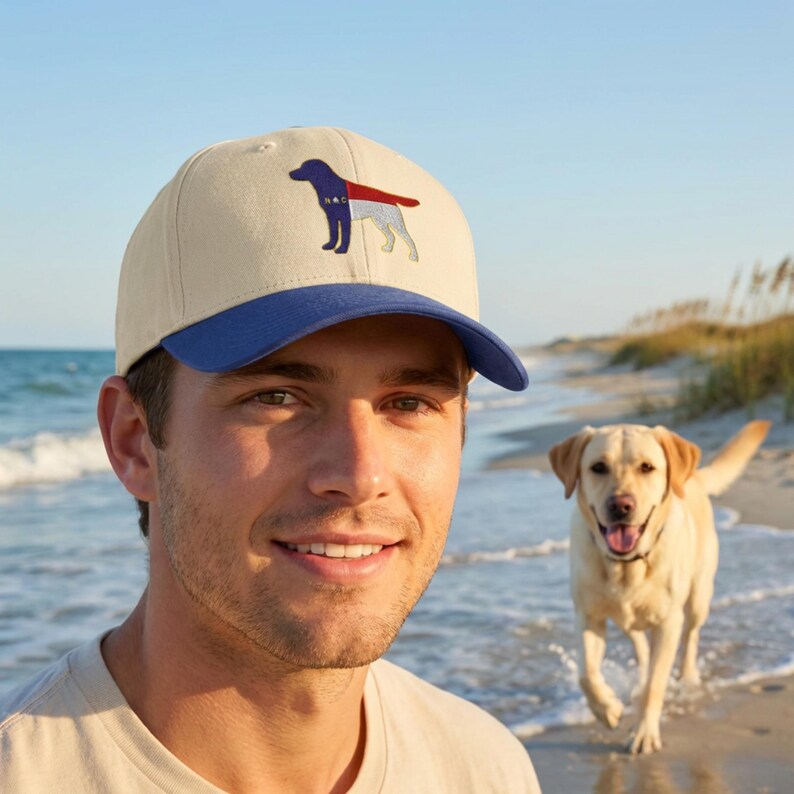 Pu&ograve; includere: Cappellino da baseball beige con visiera blu, con un disegno di cane ricamato in rosso, bianco e blu. Indossato da una persona su una spiaggia, con un cane Labrador Retriever giallo sullo sfondo.