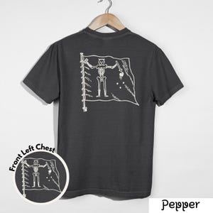 May include: Dark gray t-shirt with a white pirate flag design on the back. The flag features a skeleton figure holding a sword and a heart. A smaller version of the design is on the front left chest. The word "Pepper" is at the bottom.