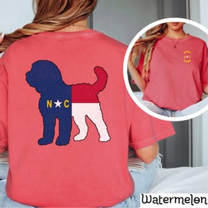 May include: A pink t-shirt with a doodle of a dog in the shape of the North Carolina flag on the back. The front of the shirt has a small North Carolina flag logo.