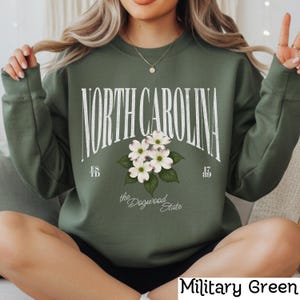 May include: Military green sweatshirt with the text "NORTH CAROLINA" in a distressed white font. Below the text is a floral design of white dogwood blossoms. The words "the Dogwood State" are printed below the flowers. The sweatshirt has the text "ESTD 1789".
