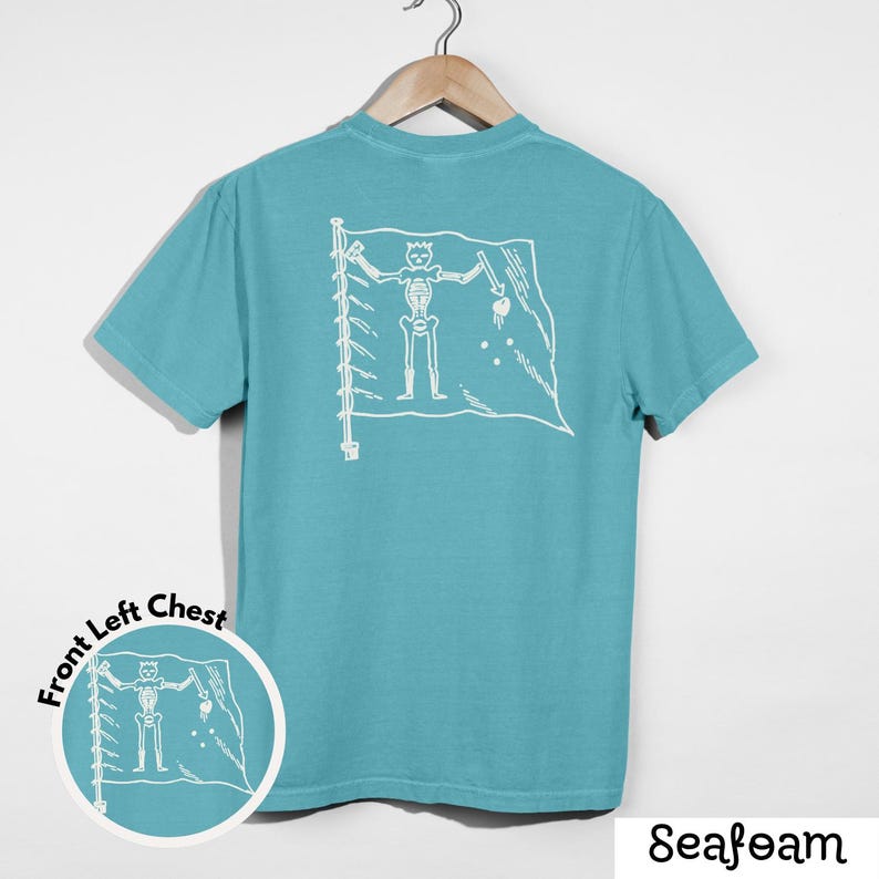 May include: Seafoam green t-shirt with a white graphic of a skeleton on a flag. The flag is on the back of the shirt. The shirt is hanging on a wooden hanger.