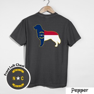 Puede incluir: Camiseta gris oscuro con la silueta de un perro que incorpora el dise&ntilde;o de la bandera del estado de Carolina del Norte. El dise&ntilde;o incluye las iniciales del estado, una estrella y las fechas 20 de mayo de 1775 y 12 de abril de 1776. La camiseta est&aacute; colgada en una percha de madera.