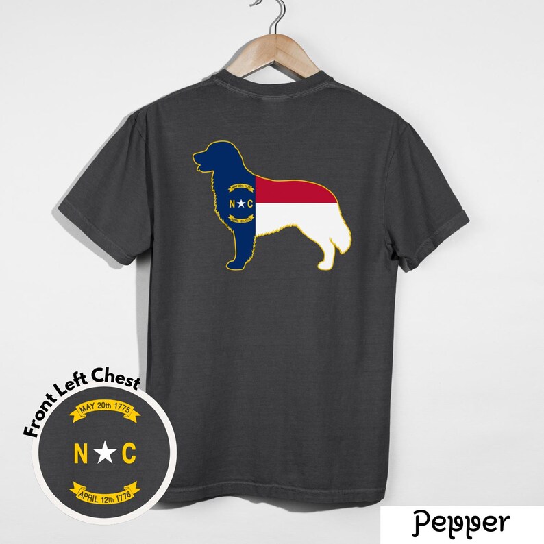 Puede incluir: Camiseta gris oscuro con la silueta de un perro que incorpora el dise&ntilde;o de la bandera del estado de Carolina del Norte. El dise&ntilde;o incluye las iniciales del estado, una estrella y las fechas 20 de mayo de 1775 y 12 de abril de 1776. La camiseta est&aacute; colgada en una percha de madera.